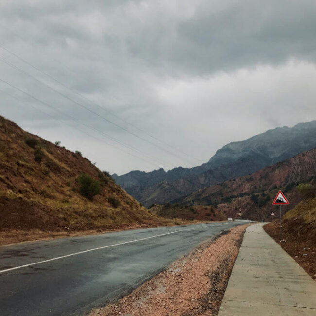 A paved road winding through a mountainous area under a cloudy sky with a steep incline road sign.