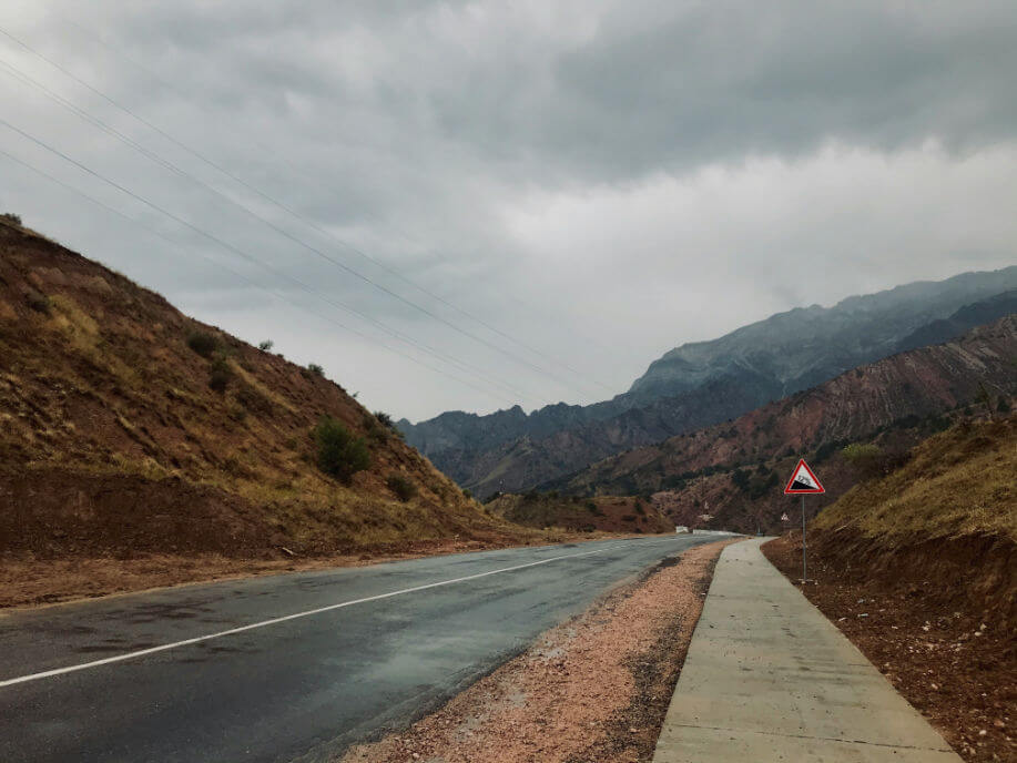 A paved road winding through a mountainous area under a cloudy sky with a steep incline road sign.